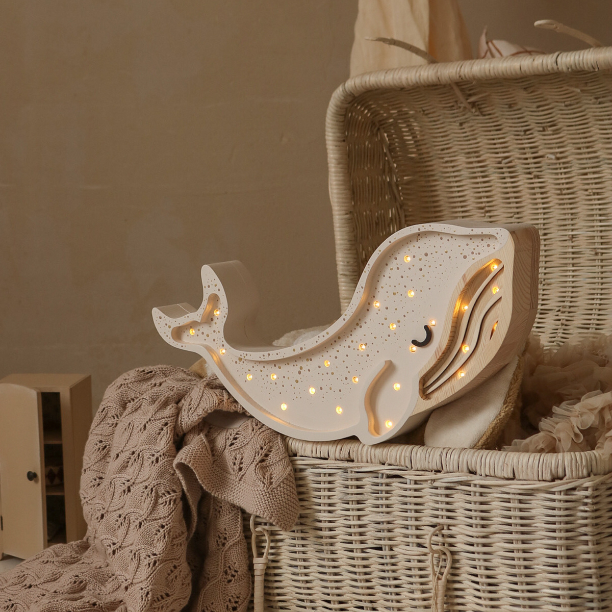 Veilleuse enfant en bois en forme de baleine, couleur crème, parsemée de petites LED allumées ; posée sur un grand panier en osier avec couverture tricotée beige ; arrière-plan doux, tons naturels.