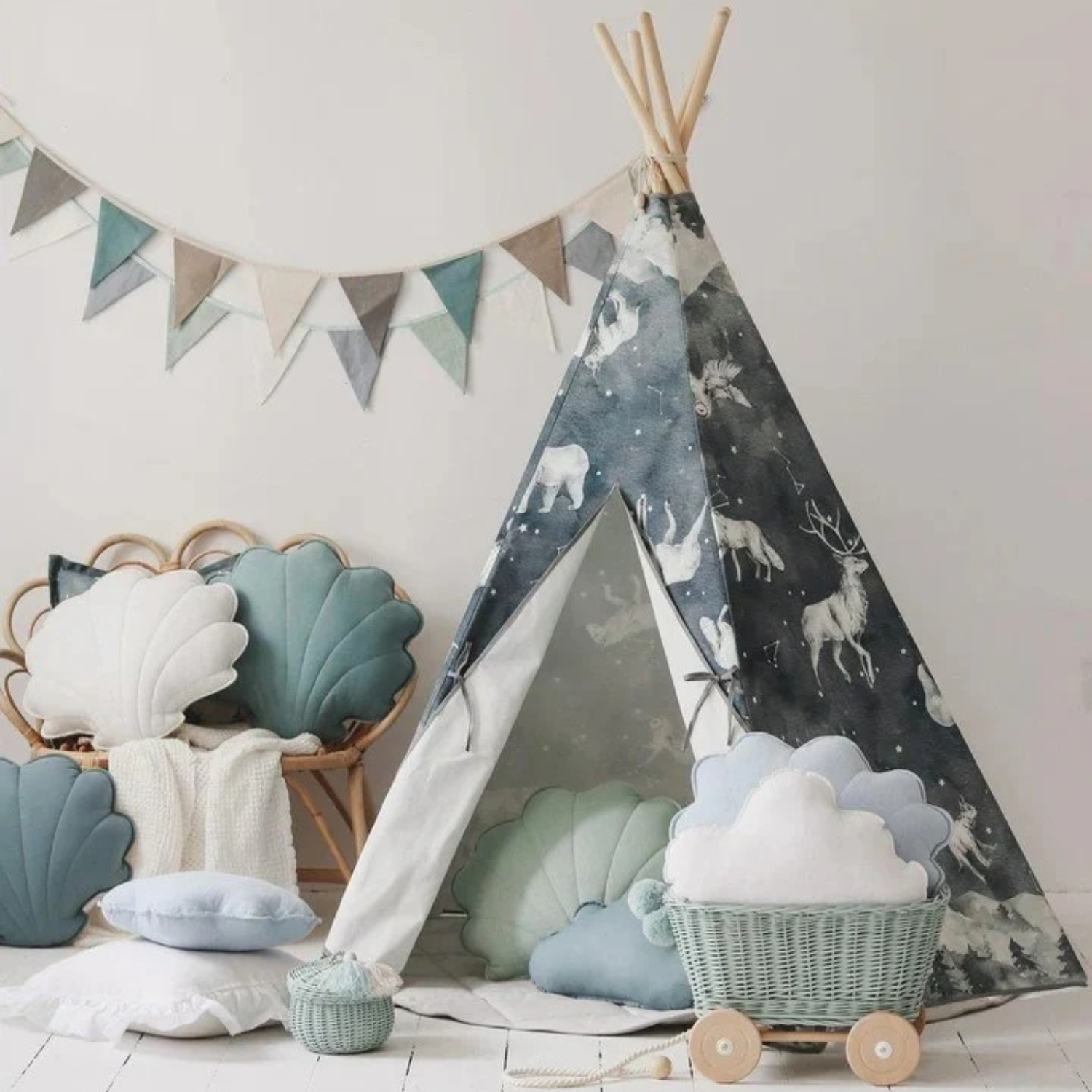 Tipi pour enfant en toile bleu nuit imprimée d’animaux (rennes, ours) avec montants en bois, entrouvert sur sol clair. Autour : coussins coquille bleu/vert, siège en rotin, panier à roulettes avec coussin nuage. Guirlande de fanions au mur.