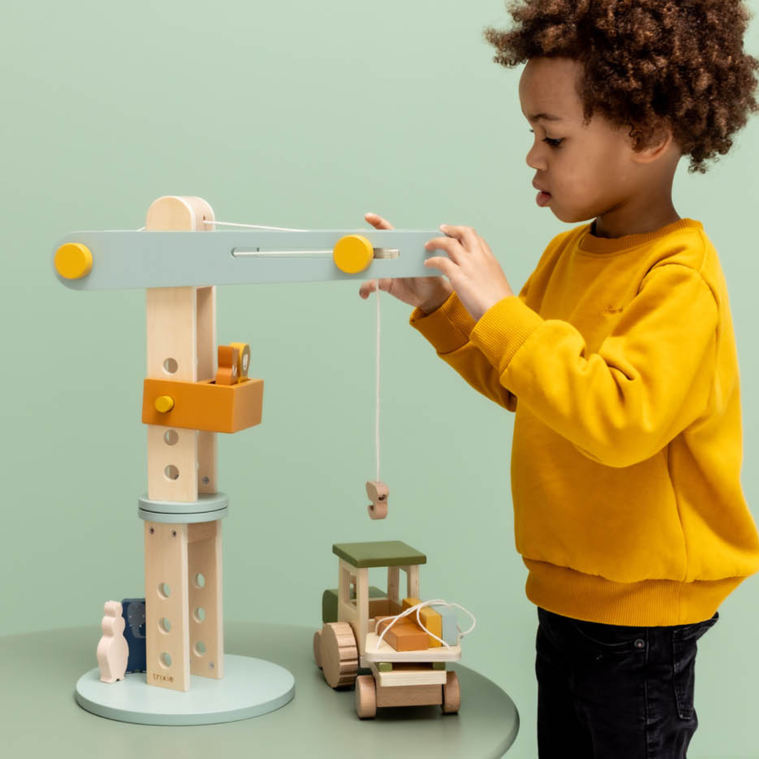 Enfant debout, sweat jaune, joue avec une grue de chantier en bois (base ronde, mât à trous, flèche vert d’eau, cabine moutarde) ; il descend un crochet à ficelle au-dessus d’un camion en bois posé sur la table ; fond vert uni.