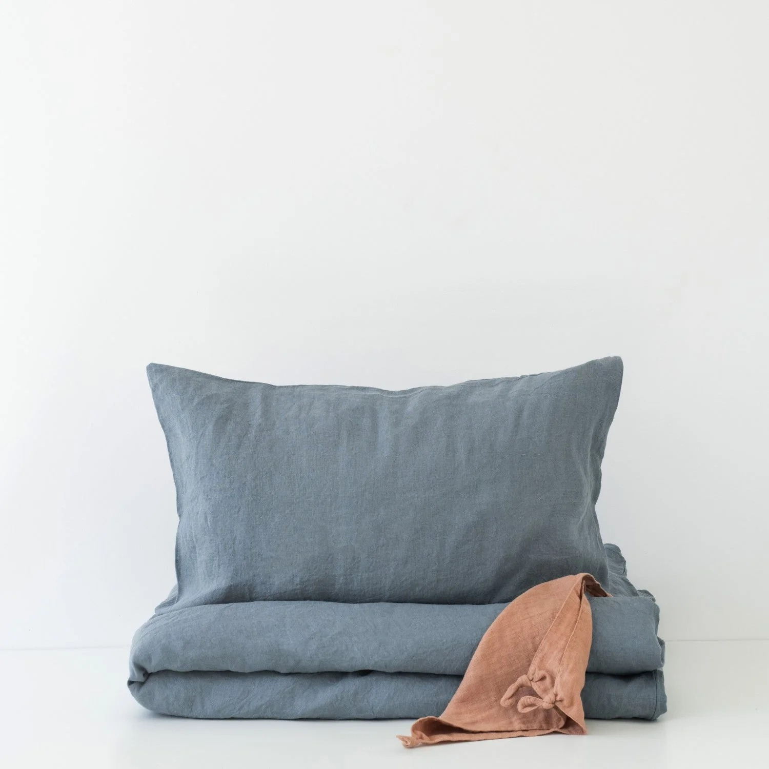 Sur fond blanc, housse de couette et taie d’oreiller en lin lavé bleu-gris, soigneusement pliées ; à droite, un petit linge terracotta avec liens noués apporte une touche de couleur.