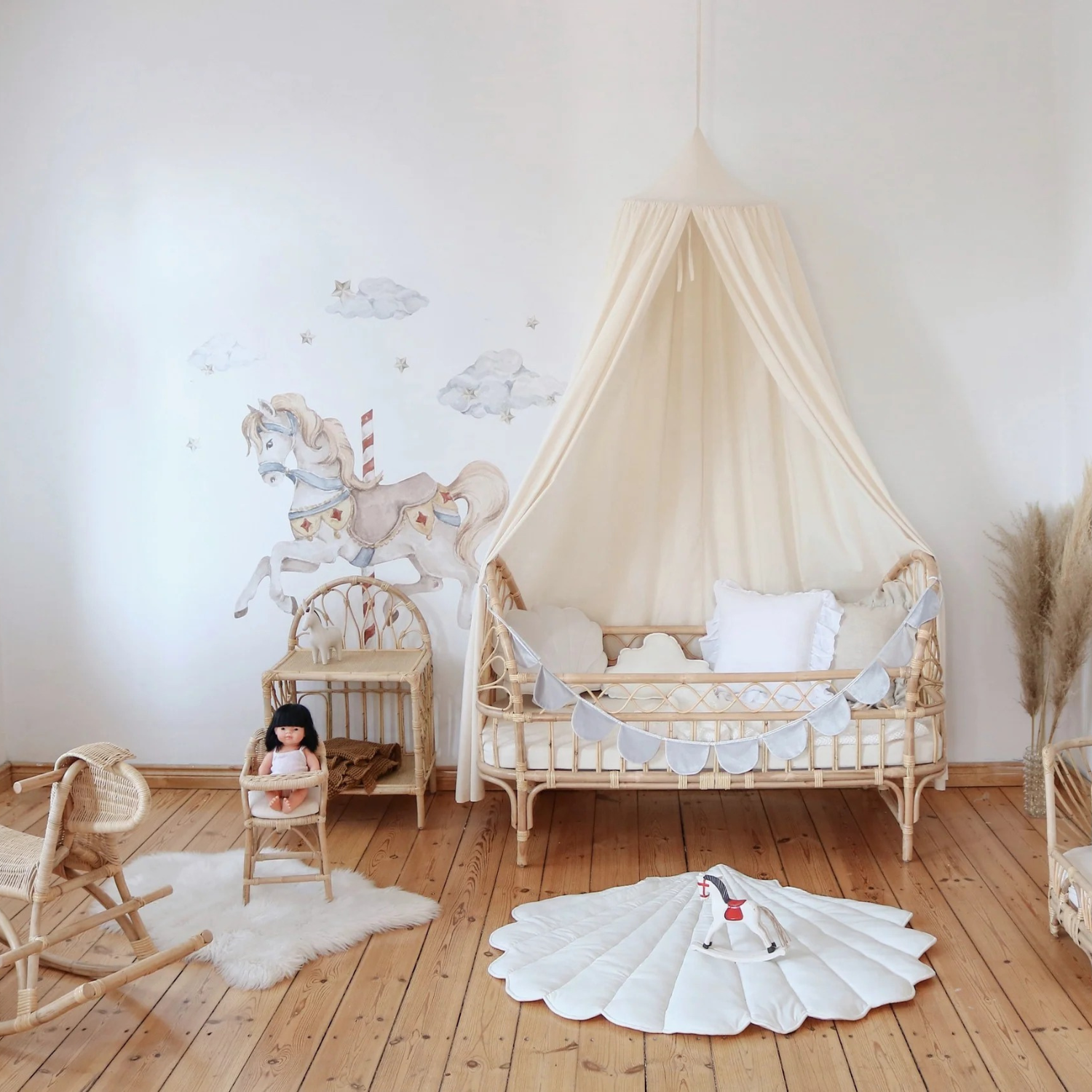 Chambre d’enfant lumineuse : lit bébé en rotin sous ciel de lit crème, guirlande gris pâle ; parquet et tapis coquille avec jouet, fauteuils en rotin, peau de mouton ; mur blanc décoré d’un cheval de manège et de nuages.
