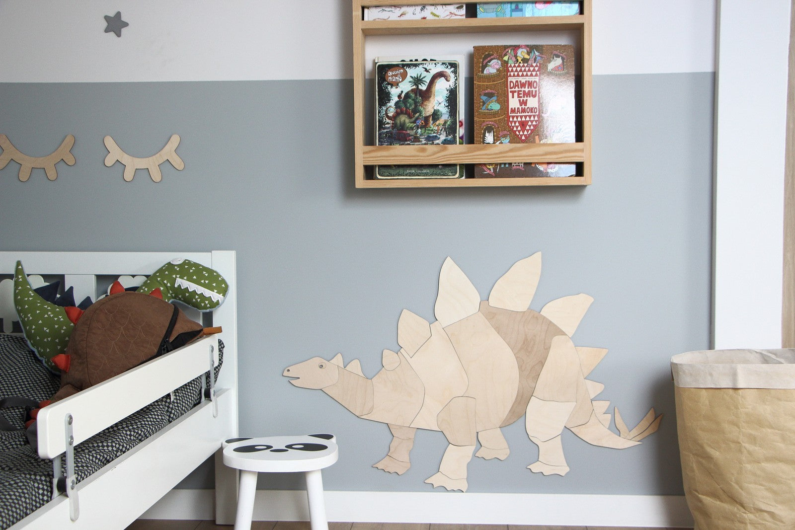 Puzzle mural en bois representant un stegosaurus stylise fixe sur un mur bicolore bleu et gris dans une chambre d’enfant. Sous cette decoration, un petit tabouret blanc est place tandis qu’a gauche, un lit enfant est orne d’un coussin dinosaure a pointes. Une etagere murale en bois expose des livres colores et une guirlande de cils decoratifs ajoute une touche ludique. Ensemble, cette scene cree un univers Montessori sur le theme des dinosaures.