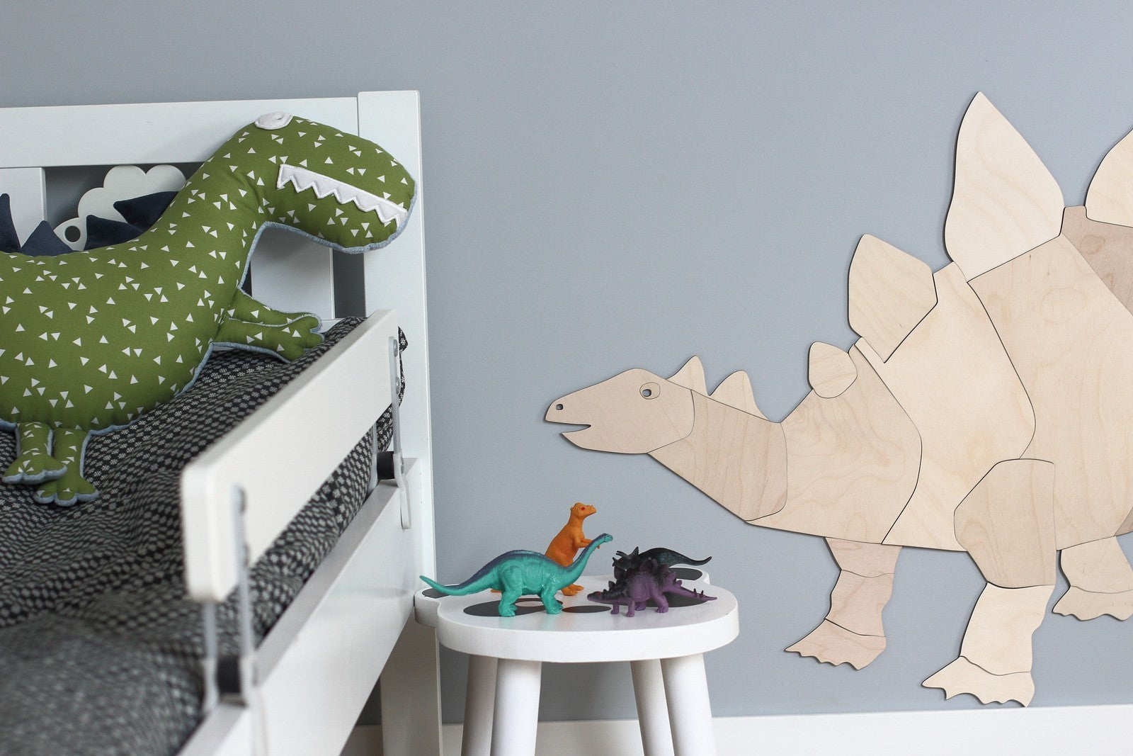 Grande silhouette de stegosaurus en bois naturel assemblee comme un puzzle et fixee sur un mur gris bleu dans une chambre d’enfant. A gauche, un lit blanc accueille une grande peluche dinosaure verte a pois, tandis qu’au premier plan une petite table ronde presente trois dinosaures jouets multicolores. Cette decoration Montessori boheme evoque un univers de dinosaures doux et naturel.