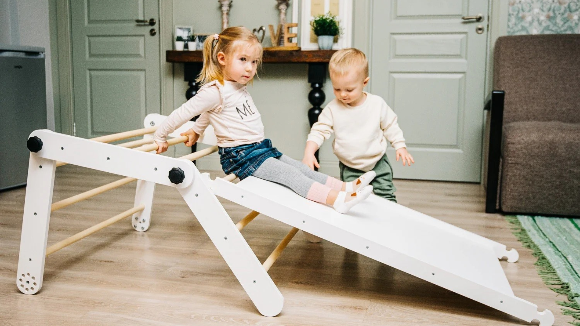 Deux jeunes enfants jouant ensemble sur un triangle de Pikler avec rampe-toboggan en bois dans un salon chaleureux, illustrant la motricité libre Montessori