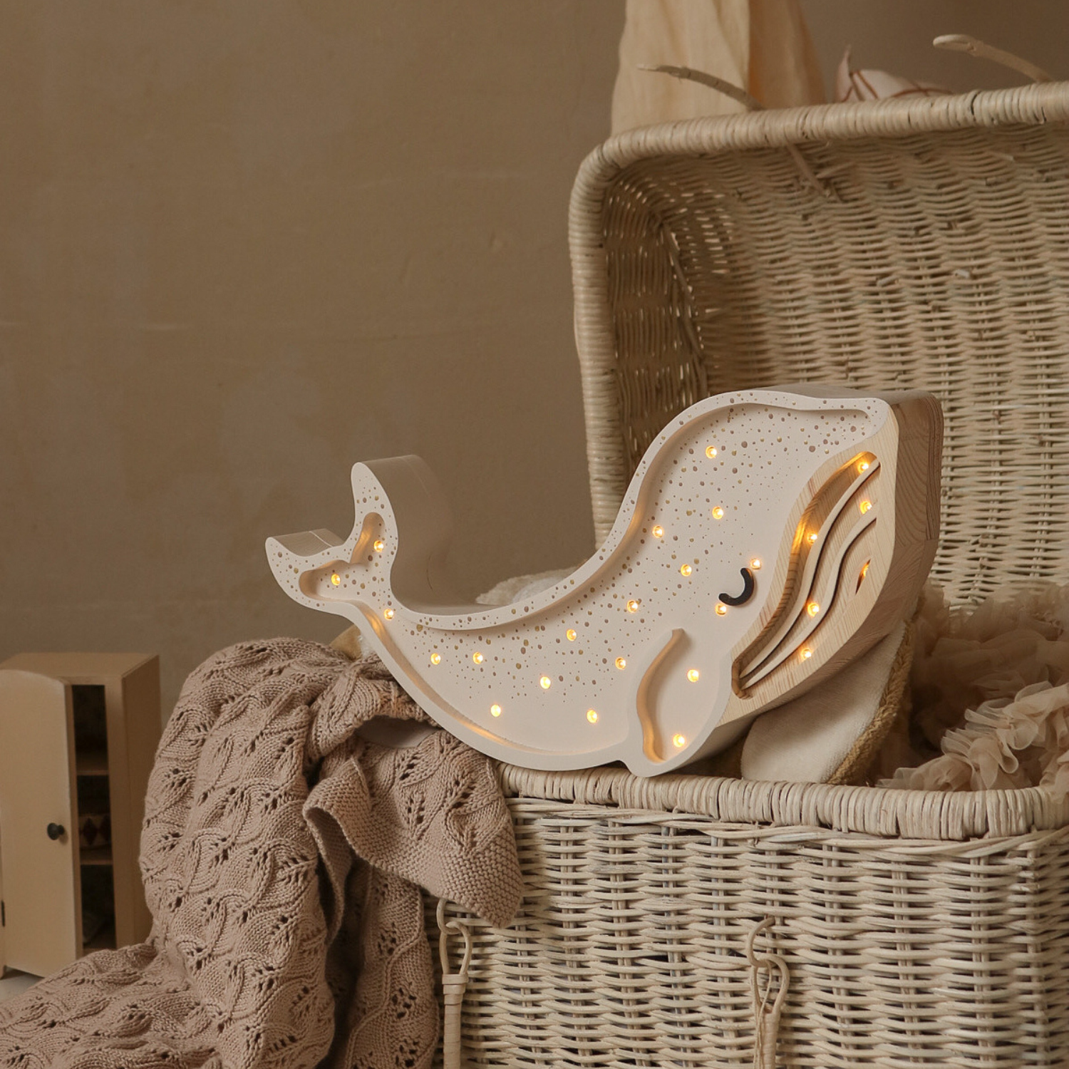 Veilleuse enfant en bois en forme de baleine, couleur crème, parsemée de petites LED allumées ; posée sur un grand panier en osier avec couverture tricotée beige ; arrière-plan doux, tons naturels.