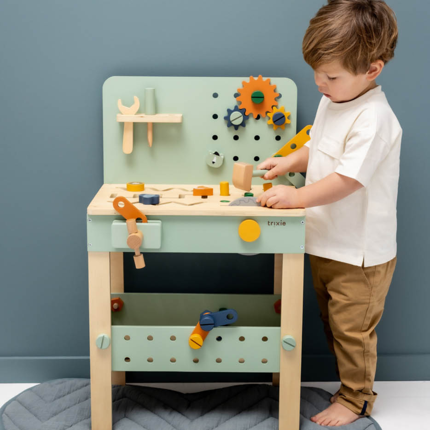 Petit garçon joue à un établi en bois vert d’eau : outils, vis et engrenages colorés sur le plateau, rangement perforé en bas ; pieds nus sur un tapis matelassé gris, mur bleu uni derrière.