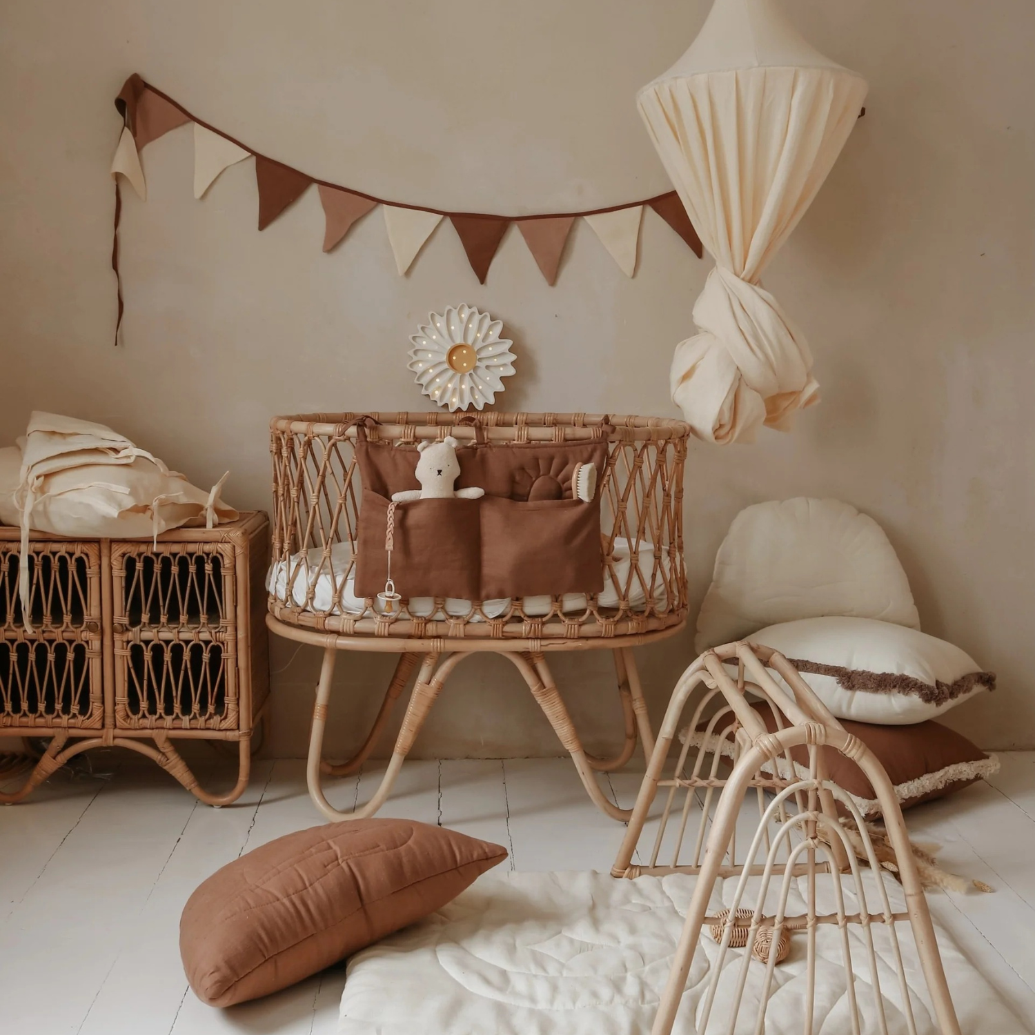 Chambre de bébé tons naturels : berceau en rotin avec organiseur brun et doudou, ciel de lit crème noué, guirlande de fanions bruns et écrus ; à droite coussins et panier, arche de jeu en rotin au sol sur tapis matelassé clair.