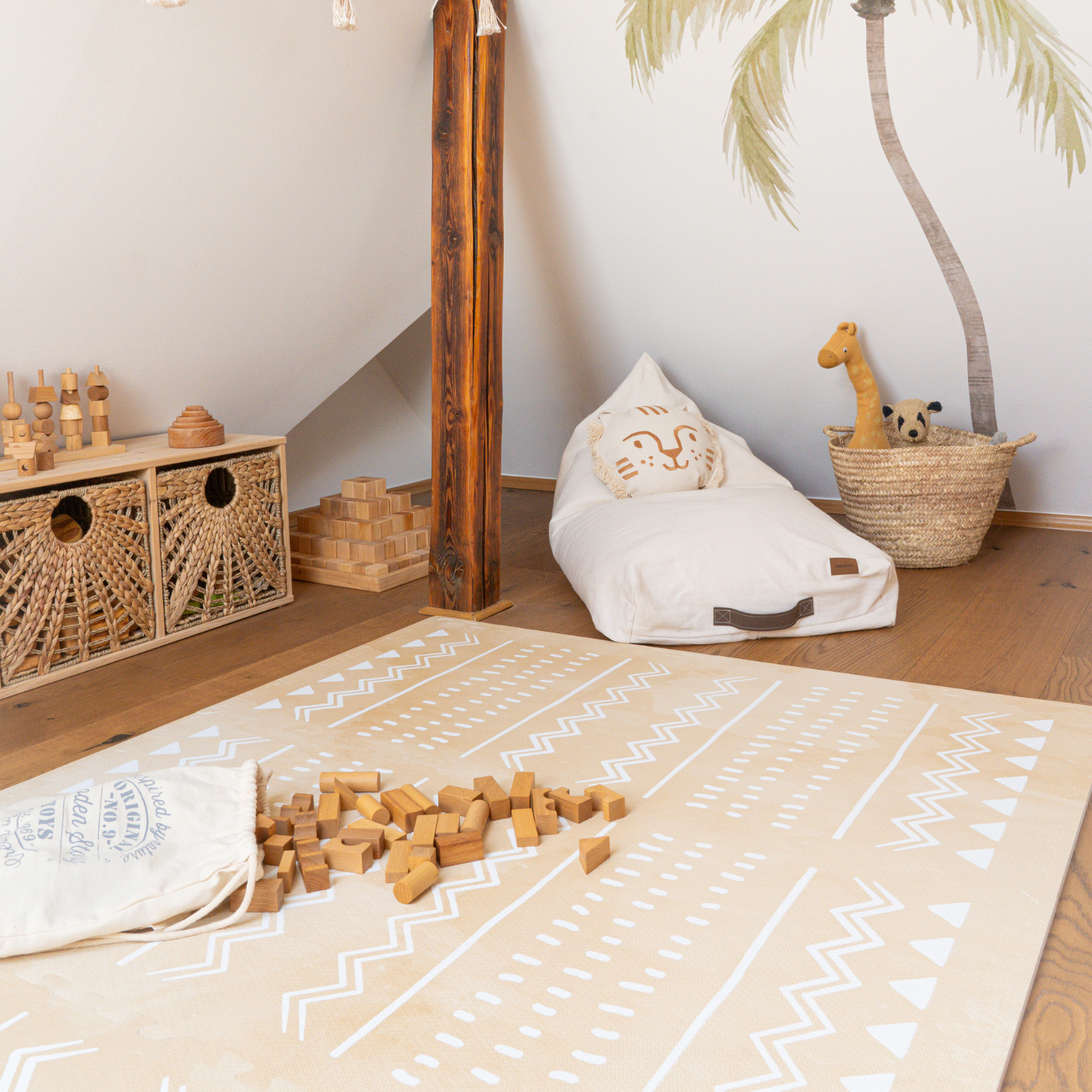 hambre d’enfant sous pente : grand tapis beige à motifs blancs, cubes en bois renversés, meuble en rotin avec jouets, pouf écru et coussin tête de chat, panier tressé avec girafe et peluche, fresque palmier au mur.