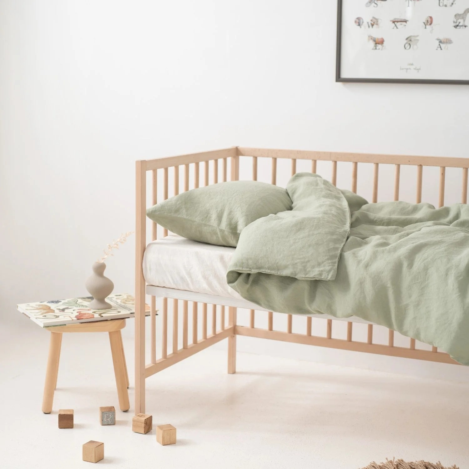 Chambre lumineuse : lit d’enfant en bois clair avec housse et oreiller en lin lavé vert sauge ; table-tabouret avec petit vase à gauche, quelques cubes en bois au sol ; mur blanc et affiche encadrée en haut à droite.