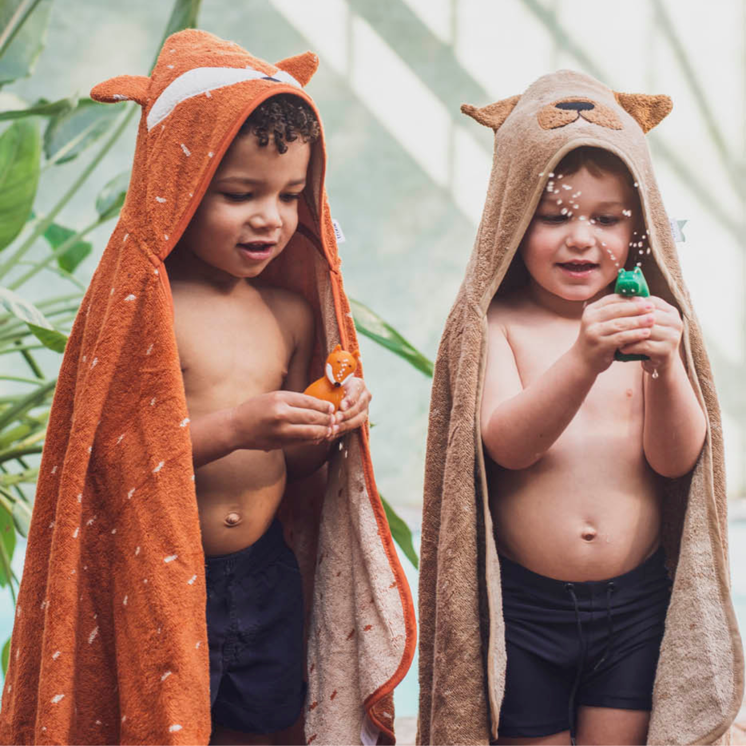 Deux enfants debout en maillot, enveloppés dans des ponchos de bain à capuche : renard orange à gauche, ours beige à droite. Ils pressent des jouets qui giclent de l’eau, gouttes visibles ; arrière-plan lumineux avec plantes.