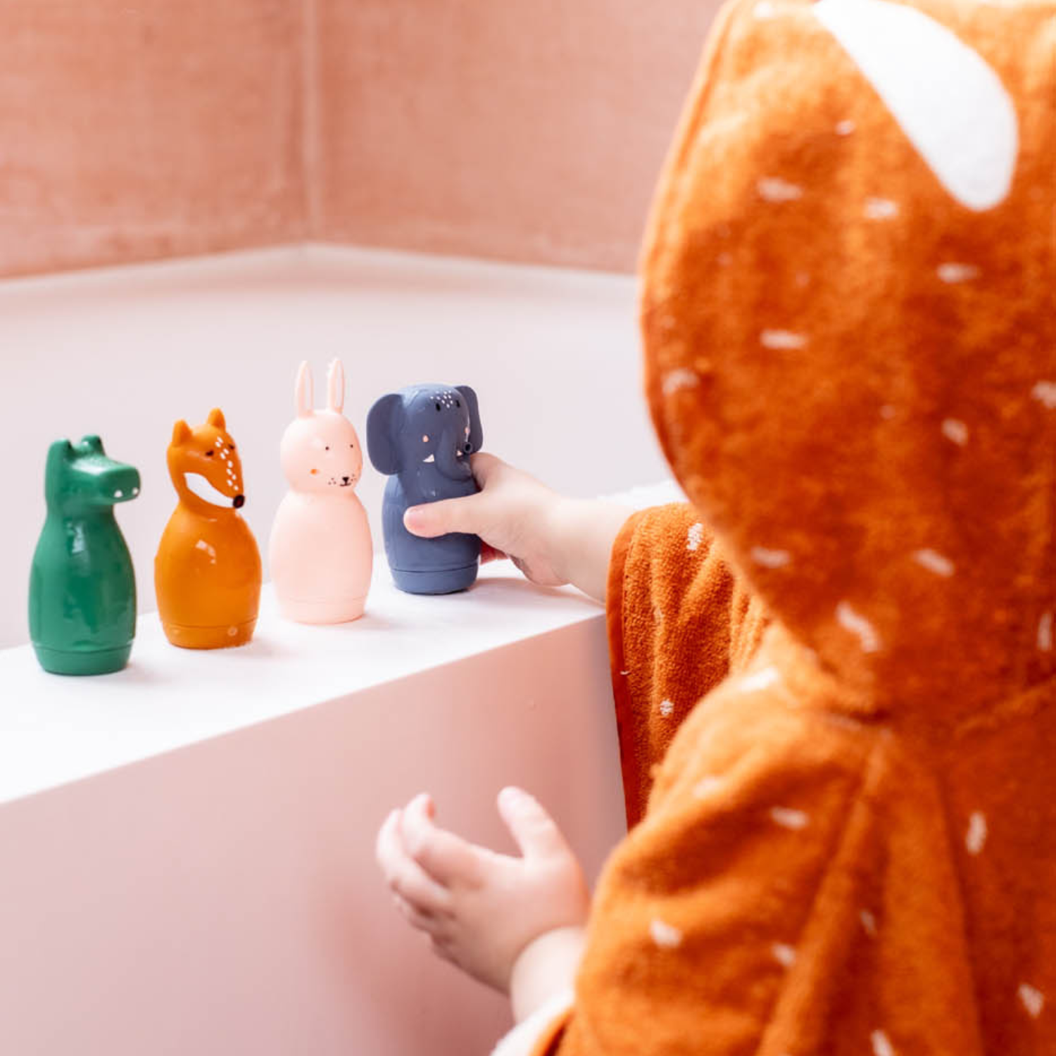 Enfant de dos en poncho de bain orange à capuche renard, près d’une baignoire. Sur le rebord, quatre jouets de bain alignés : crocodile vert, renard ocre, lapin rose pâle et éléphant bleu. Lumière douce, carrelage rosé.