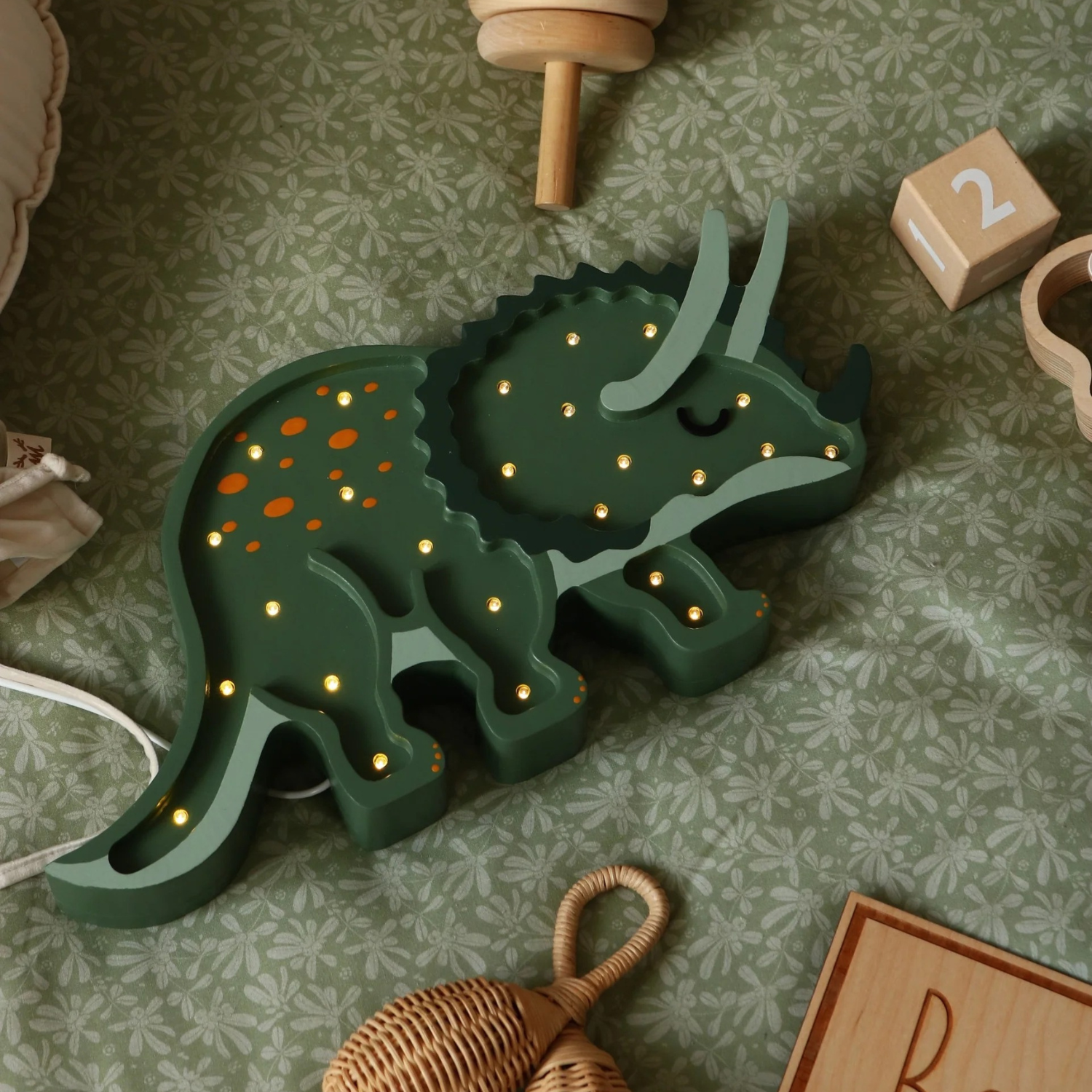 Veilleuse Little Lights en bois, tricératops vert parsemé de petites LED allumées et de points orange ; posée sur tissu vert à motifs. Autour : jouets en bois dont hochet tressé et cube chiffré ; câble blanc visible à gauche.