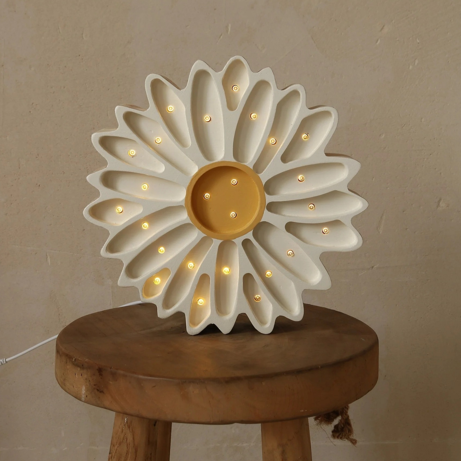 Veilleuse enfant en forme de marguerite, pétales crème et cœur jaune, parsemée de petites LED allumées ; posée sur un tabouret en bois rond, fond mur beige, câble blanc visible à gauche.