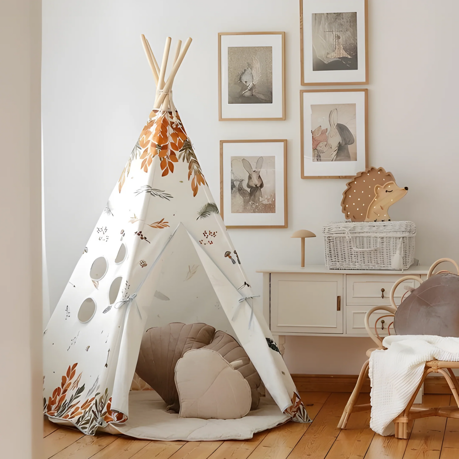Tipi pour enfant en toile crème décorée de feuilles d’automne, entrouvert sur un tapis rond beige avec deux coussins en forme de cœur ; chambre lumineuse au sol en bois, cadres au mur et panier en osier à droite.