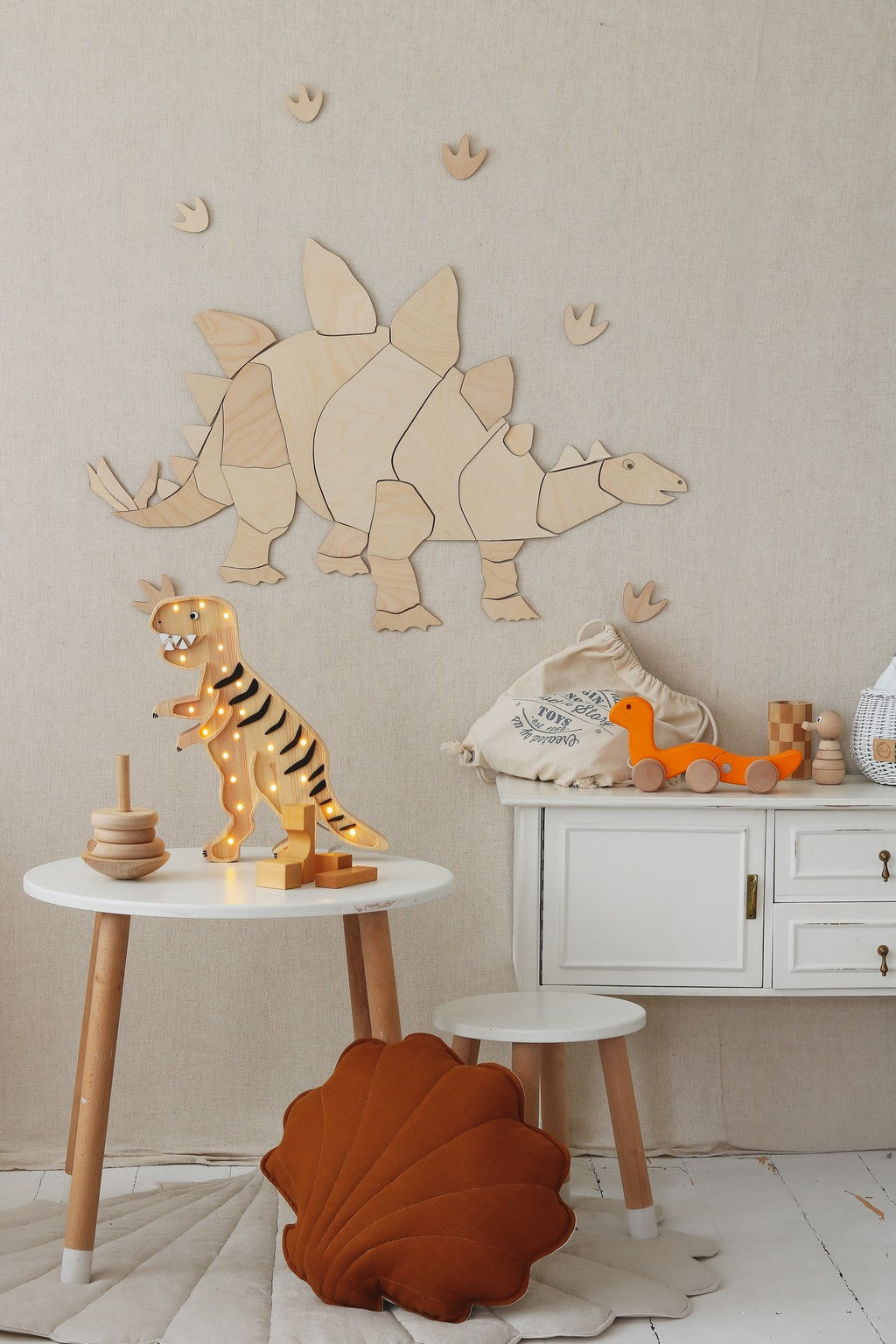 Puzzle mural en bois representant un stegosaurus stylise et ses empreintes fixe sur un mur beige. Devant, une table ronde accueille un jouet dinosaure lumineux en bois et un anneau d’empilement; un meuble blanc supporte une petite lampe dinosaure orange. Un coussin coquillage rouille sur le sol complete cette decoration Montessori boheme pour chambre d’enfant sur le theme des dinosaures.