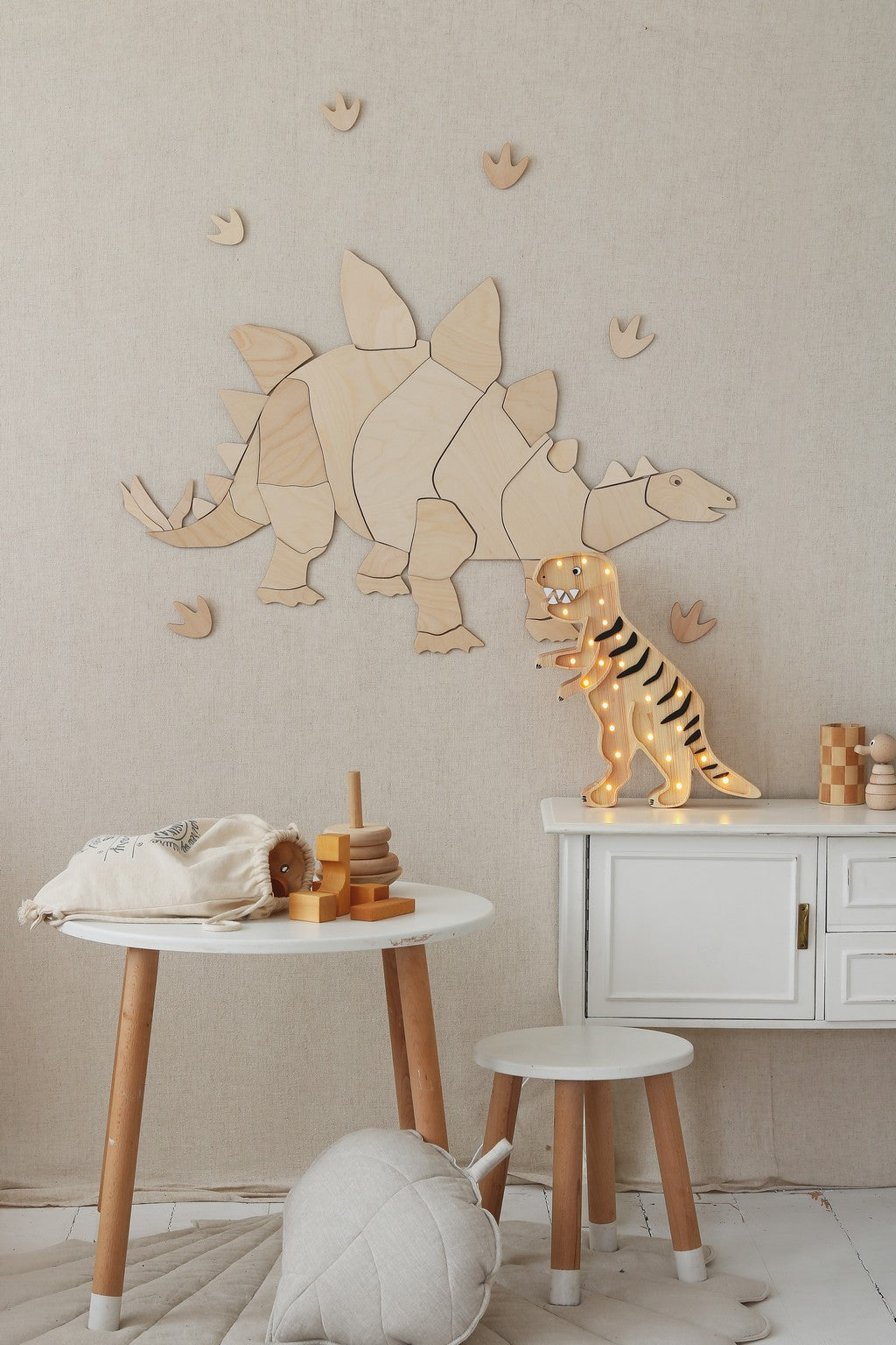 Puzzle mural en bois representant un dinosaure stegosaurus stylise et ses empreintes, fixe sur un mur en tissu beige; en dessous, une table ronde blanche avec jouet d'equilibre en bois et doudou, ainsi qu'une console et un tabouret surmontes d'une figurine de dinosaure orange; decor naturel Montessori pour chambre d'enfant sur le theme des dinosaures