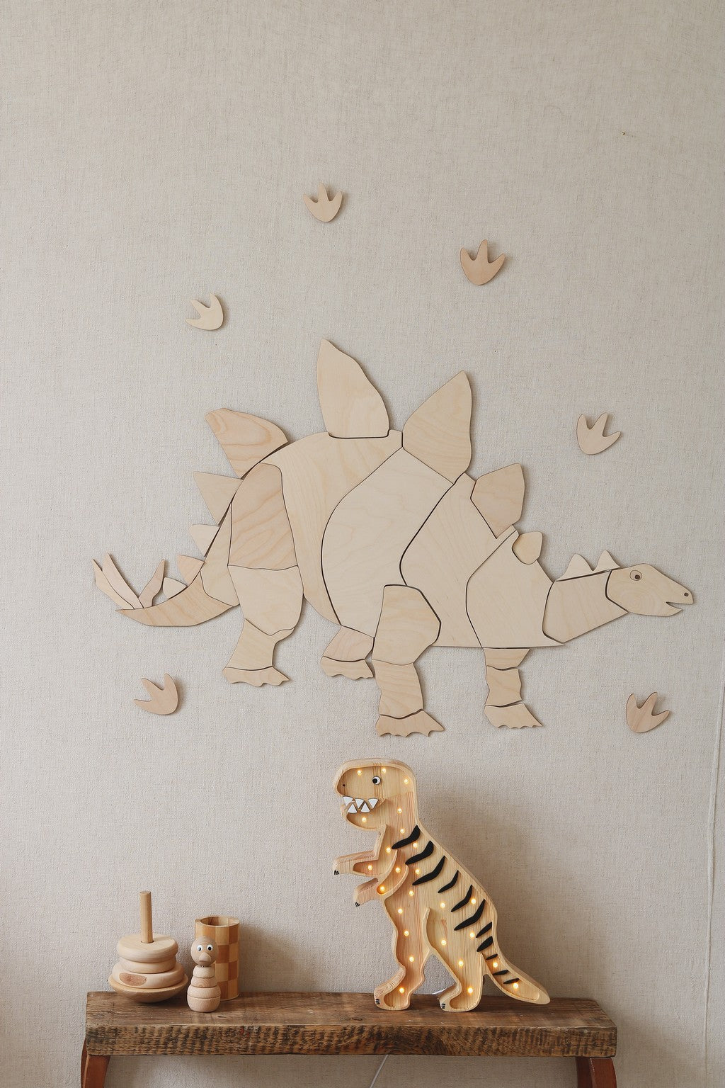 Décoration murale en bois représentant un stégosaurus dans une chambre d'enfant. Le dinosaure en bois naturel est composé de pièces de puzzle fixées sur un mur beige avec des empreintes de pas de dinosaures tout autour. Sur la table en bois située en dessous, un jouet dinosaure articulé en bois et un jeu d'anneaux empilables complètent la scène. Décoration Montessori et naturelle, thème dinosaure, idéale pour la chambre d'enfant.