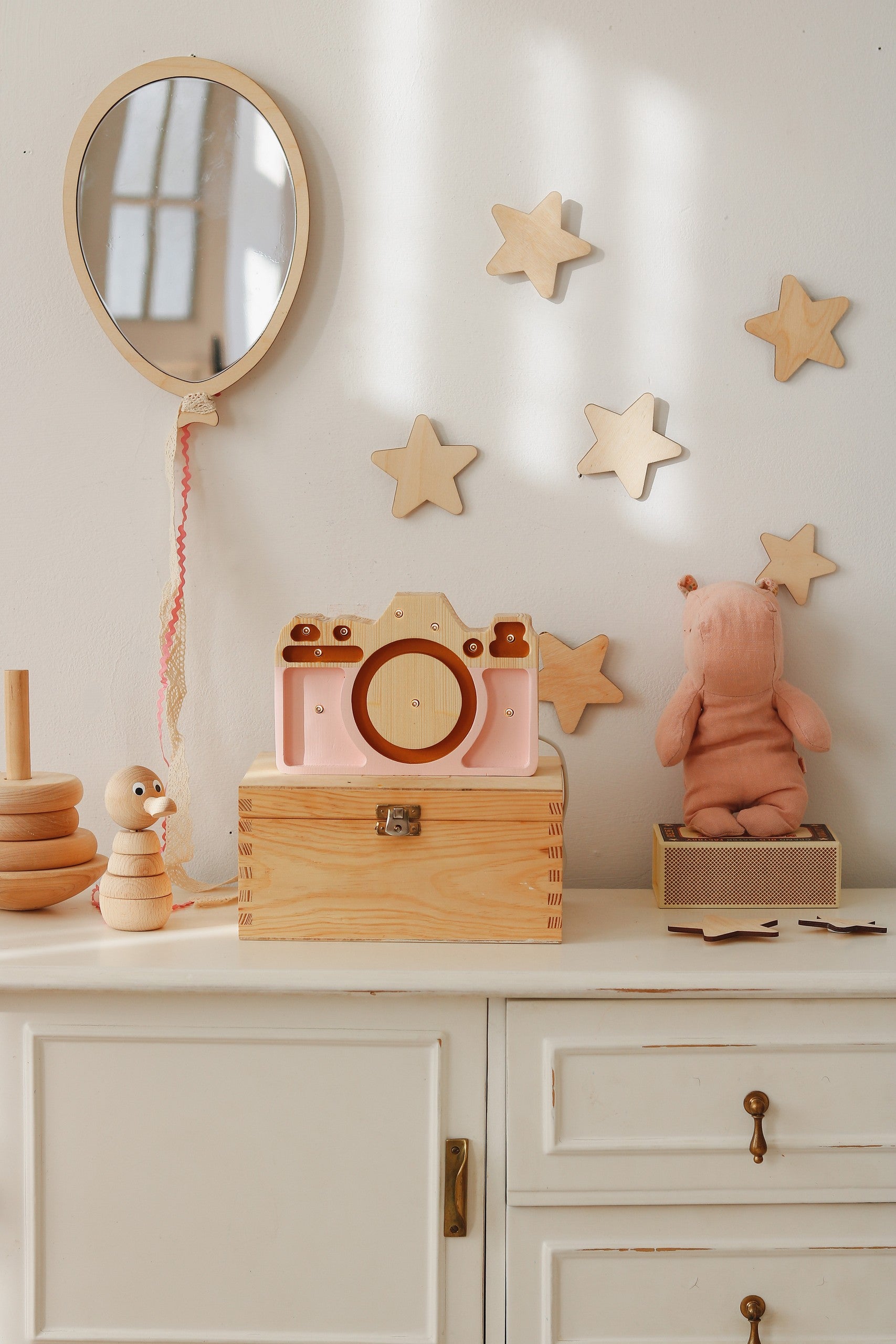 Scène décorative dans une chambre d'enfant au style bohème et naturel. Un miroir en forme de ballon de baudruche fabriqué en bois et acrylique est suspendu au mur blanc par une cordelette. Autour du miroir, des stickers étoiles créent une ambiance cosy. Sur la commode blanche en bois se trouvent un appareil photo jouet en bois, un miroir rond encastré dans une boîte, une tirelire maison, un escargot en bois, un panier tressé et une peluche rose en forme de lapin. Décoration Montessori pour chambre de bébé o