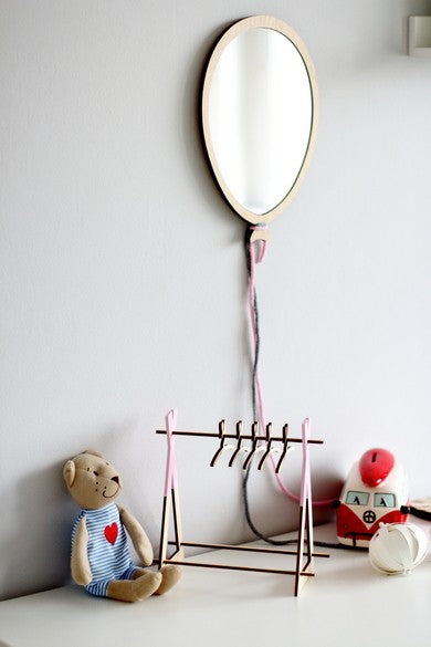 Miroir en forme de ballon suspendu au mur gris par une corde rose, fabriqué en bois et acrylique. Sous le miroir, un petit portant en bois sert d'étagère. On y voit un ours en peluche portant une salopette à rayures bleues avec un coeur rouge, une voiture jouet vintage rouge et blanche, un chapeau beige et des cintres. L'ensemble offre une ambiance minimaliste et scandinave, parfait pour la décoration Montessori et boho d'une chambre d'enfant.