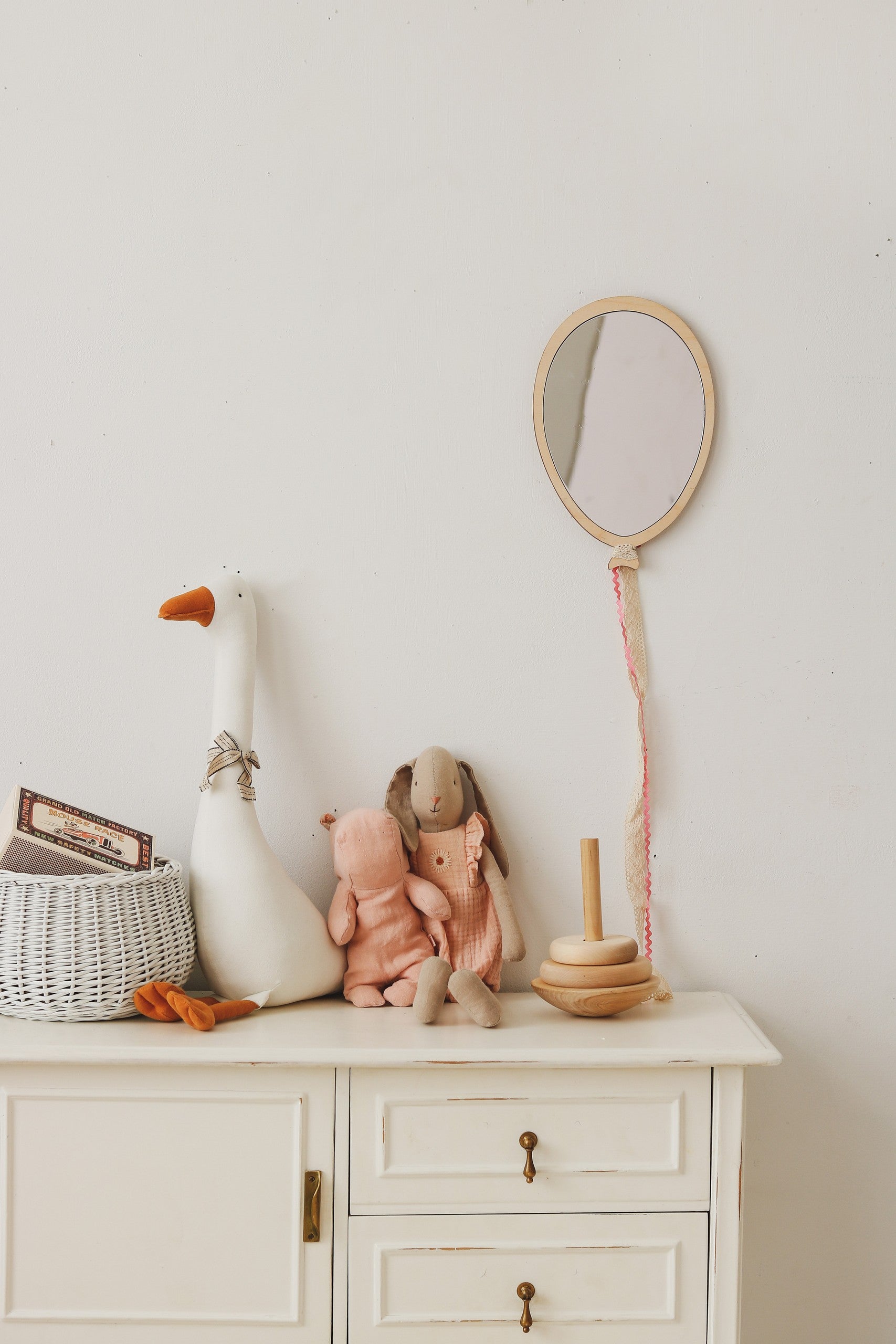 Ambiance douce et naturelle dans une chambre d'enfant. Un miroir en forme de ballon fabriqué en bois et acrylique est suspendu au mur blanc par une cordelette. Sur la commode blanche se tiennent une grande peluche d'oie blanche avec foulard, deux doudous en tissu rose et beige, un jouet empilable en anneaux de bois, un panier tressé avec couvercle et un petit cochon en bois. La décoration au style boho et Montessori combine des tons neutres et matériaux naturels, idéal pour une chambre de bébé ou d'enfant.