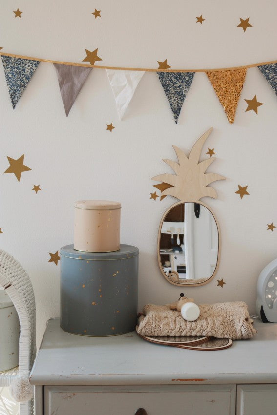 Guirlande de fanions et étoiles dorées décorant une chambre d’enfant avec miroir en forme d’ananas en bois et acrylique au mur. Sur la commode vintage se trouvent une boîte cylindrique bleue étoilée, un pot beige et un panier en osier rempli de linge, créant une ambiance tropicale et Montessori.