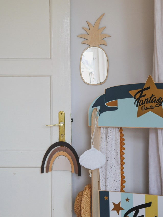 Composition décorative pour porte de chambre d’enfant mettant en scène un miroir en forme d’ananas en bois et acrylique, un arc‑en‑ciel en bois suspendu à la poignée dorée, un panneau en bois aux couleurs pastel marqué « Fantaisie », un nuage en laine crocheté, une guirlande de perles et un sac en osier posé à terre. Ce décor tropical et bohème, aux teintes douces, convient à une chambre Montessori sur le thème de l’arc‑en‑ciel et de l’ananas.
