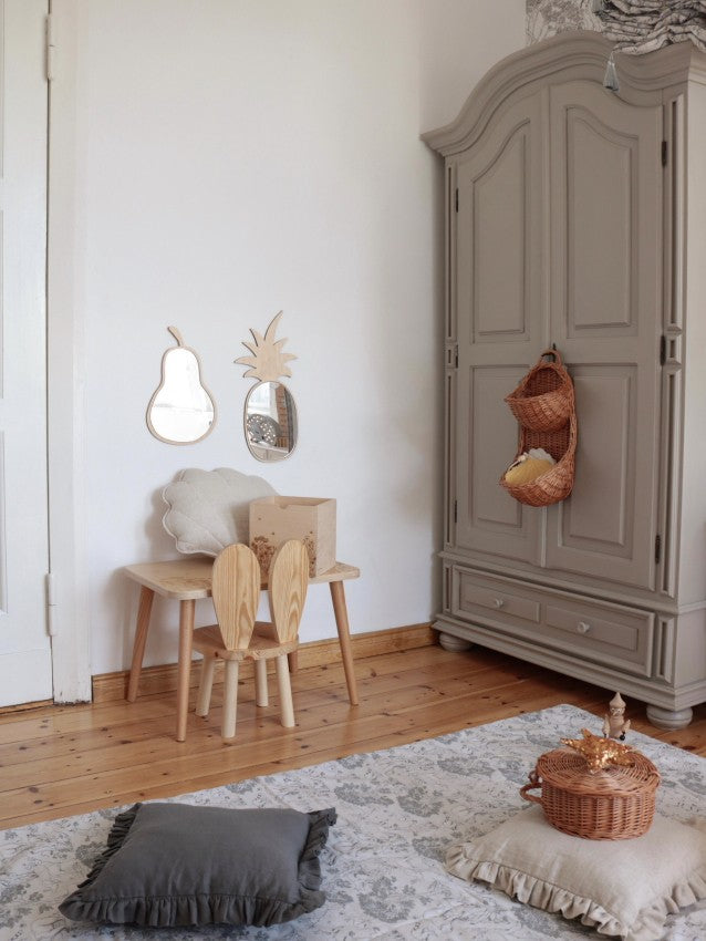Ambiance épurée et bohème dans une chambre d’enfant avec un miroir en forme d’ananas en bois et acrylique et un miroir en forme de goutte suspendus au mur blanc à côté d’une grande armoire beige. Le coin jeu est aménagé avec un bureau et des chaises en bois clair aux dossiers en forme de feuilles, un coussin en fausse fourrure et un panier en osier tressé. Un chapeau et un sac en paille pendent à côté. Un tapis persan pastel et un coussin gris anthracite complètent cette décoration Montessori tropicale.