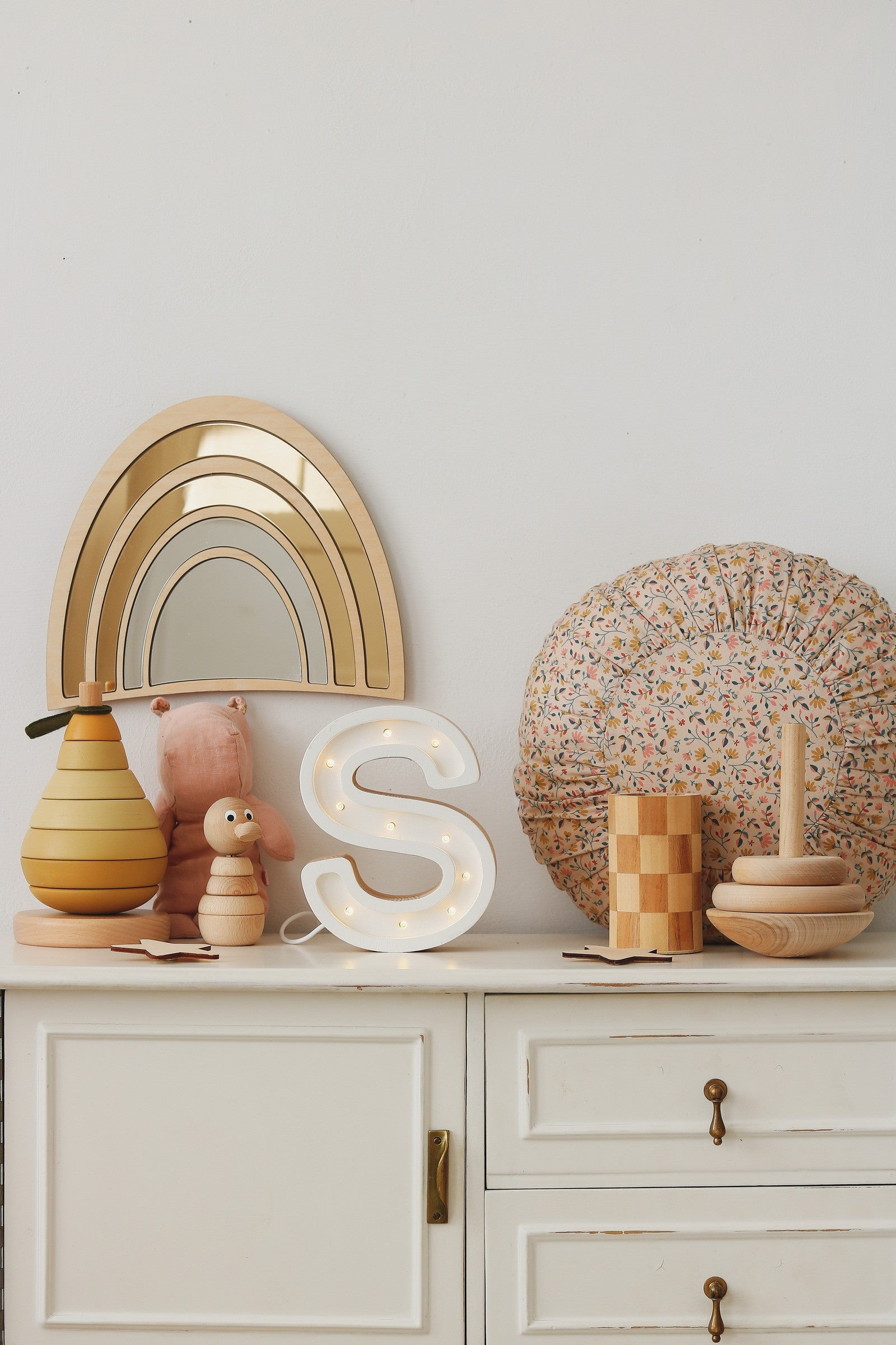 Composition décorative bohème pour chambre d’enfant avec un miroir arc‑en‑ciel doré en bois et acrylique appuyé contre un mur, une peluche chat rose pastel, une grande housse de coussin ronde à motif liberty, une lettre lumineuse S, un pot en bois à damier et un vase rond. Cette scène chaleureuse, aux matières naturelles et couleurs douces, convient à une chambre Montessori, décoration arc‑en‑ciel et ambiance bébé neutre.