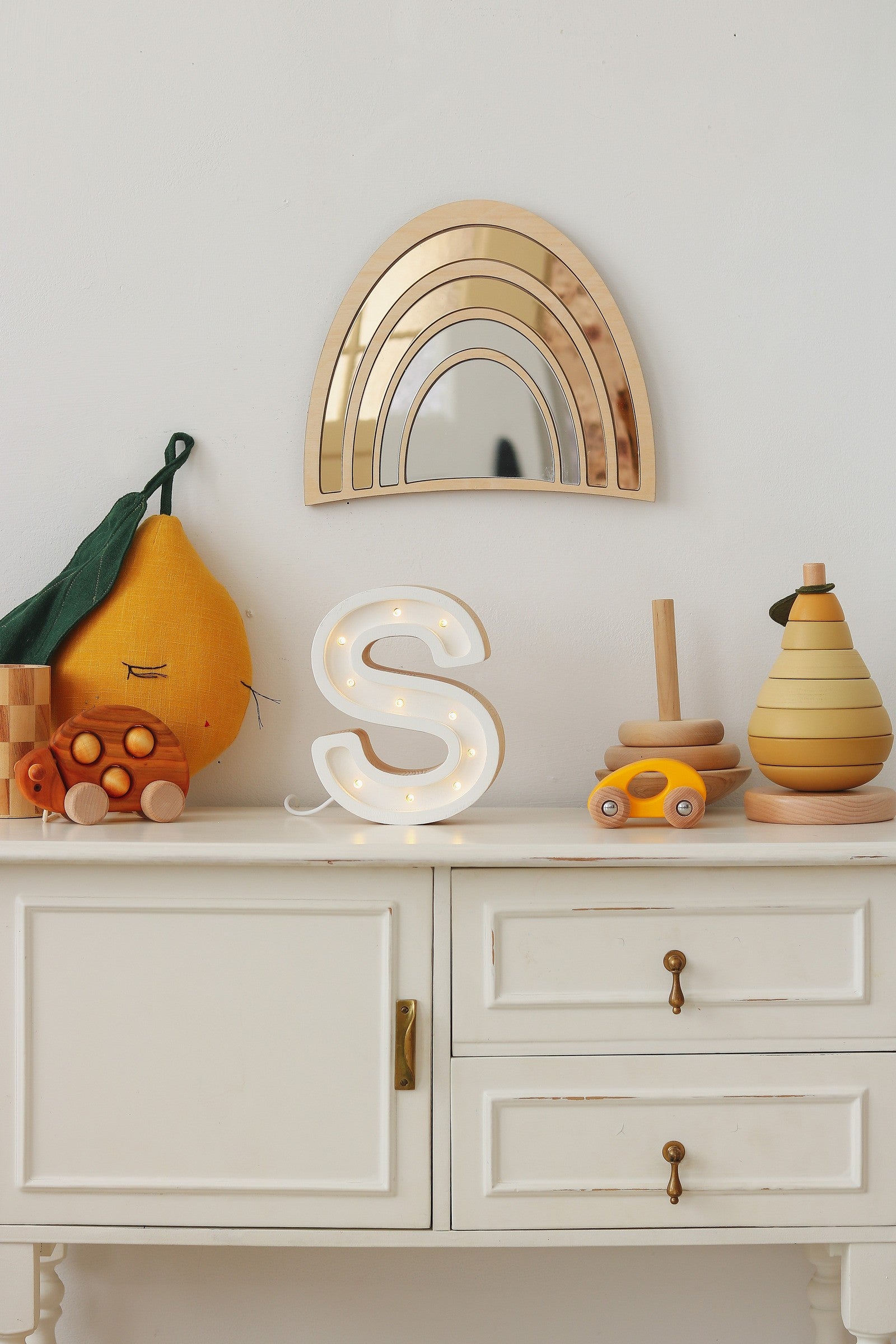 Scène de chambre d’enfant bohème avec miroir arc-en-ciel en bois et acrylique accroché au mur au-dessus d’une commode blanche vintage ; sur la commode se trouvent une lettre décorative lumineuse en forme de S, des peluches représentant des fruits orange et poire, et un vase amphore en bois. Cette décoration naturelle et chaleureuse est idéale pour une chambre Montessori, un thème arc‑en‑ciel et une ambiance neutre pour bébé.