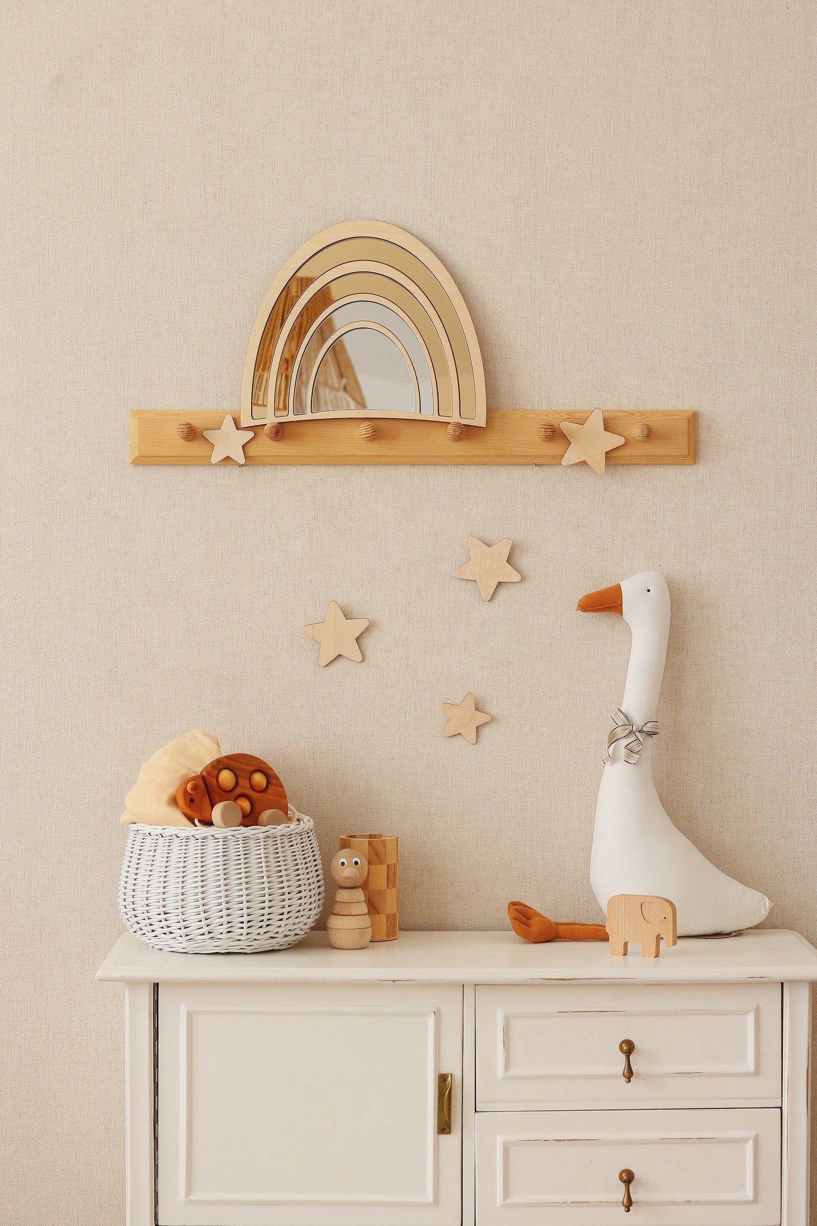 Miroir arc-en-ciel en acrylique et bois monté sur étagère ornée d'étoiles, décoration murale pour chambre d'enfant, accompagné d'un panier tressé avec jouets en bois, d'une peluche oie blanche et de petites étoiles sur un mur beige, style naturel Montessori pour une chambre de bébé chaleureuse avec mots-clés miroir arc-en-ciel, bois et décor Montessori.