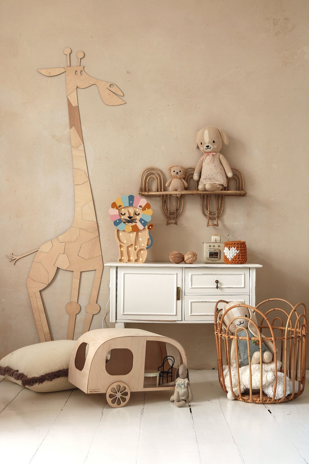 Deco murale en bois representant une grande girafe stylisee facon puzzle appuyee contre un mur beige dans une chambre d’enfant. Devant la girafe se trouve une commode blanche avec etagere suspendue garnie de peluches et d’une figurine de lion colore. Au sol, un gros coussin, une caravane jouet en bois, une poussette en rotin et une poupee en tissu composent une mise en scene safari boheme et Montessori.