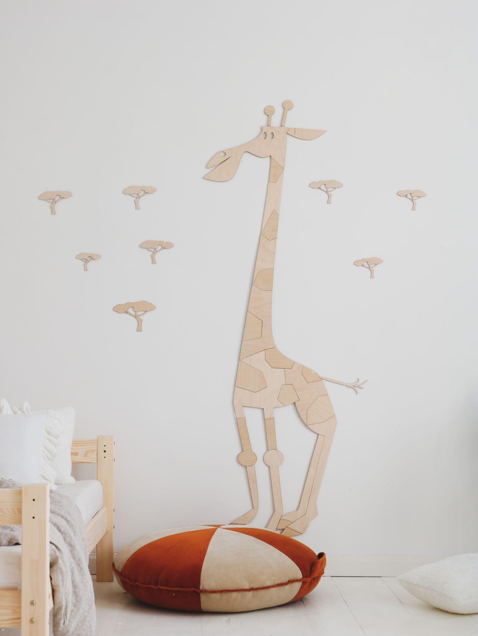 Grande girafe stylisee en bois naturel composee de pieces puzzle fixee au mur d'une chambre d'enfant au theme safari avec petits arbres de savane, lit en bois clair et coussin rond terracotta; decoration murale Montessori boho