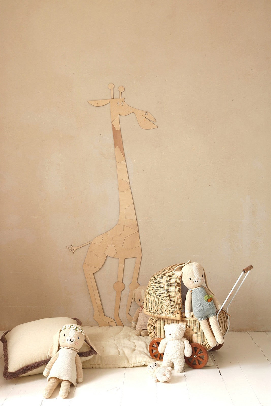 Decor mural en bois representant une grande girafe stylisee composee de pieces puzzle fixees sur un mur beige dans une chambre d’enfant au theme safari. Au sol, des coussins en lin beige, un panier en osier rempli de peluches et une poupée en tissu apportent une touche boheme et naturelle a la decoration Montessori.