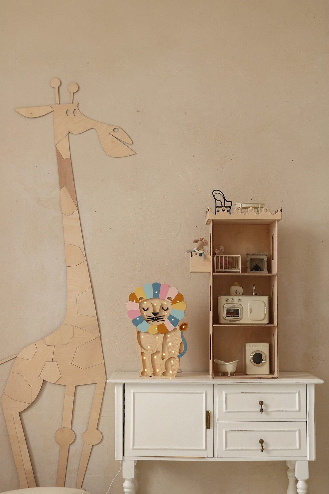 Grande girafe en bois naturel facon puzzle appuyee contre un mur beige, a cote d'une commode blanche sur laquelle se trouvent une figurine de lion coloree, une maison miniature et des jouets vintage; decoration safari boho pour chambre Montessori